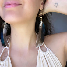 Load image into Gallery viewer, Woman wearing a Fauna eucalyptus and organic cotton maxi dress and cruelty free feather earrings made from ethically sourced duck feathers. Beautiful colors found in each unique feather, from white and gray to iridescent hues of purples, greens, and blues. Each earring also features antique bronze fold over clasps, raw brass rings in different sizes, and raw brass french hook ear wires.
