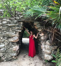 Load image into Gallery viewer, A woman standing in Tulum, Mexico with stone arches and greenery in the background, wearing a Fauna eucalyptus and organic cotton red maxi dress with keyhole details at the shoulders and mid bust.
